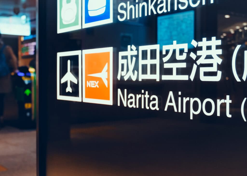 A station sign for Narita Airport with airplane icons and the N'EX logo at a Tokyo railway station