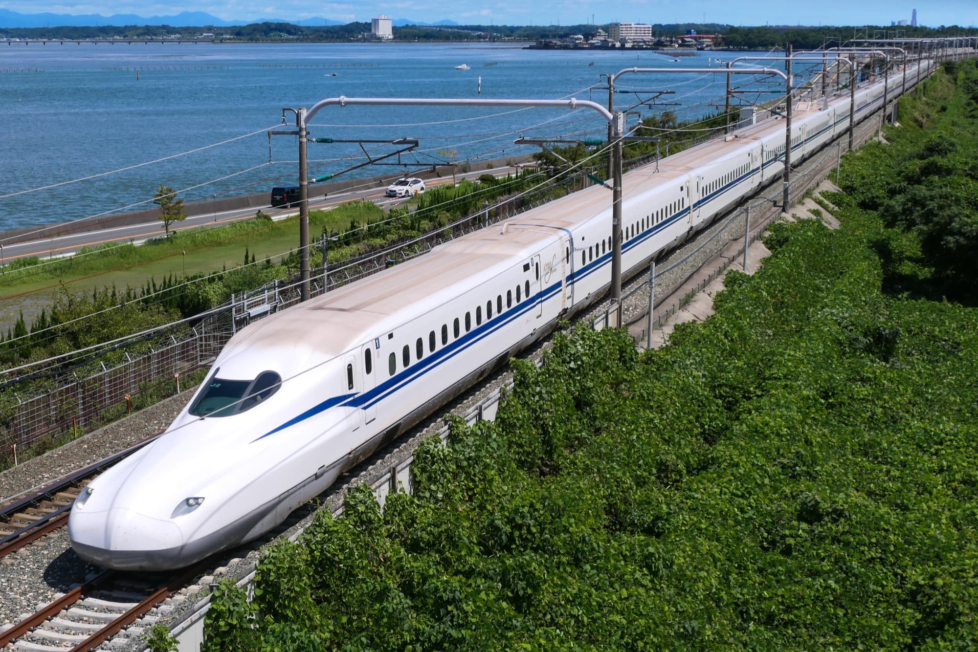 A Japanese Shinkansen train running along the coast next to a calm blue bay
