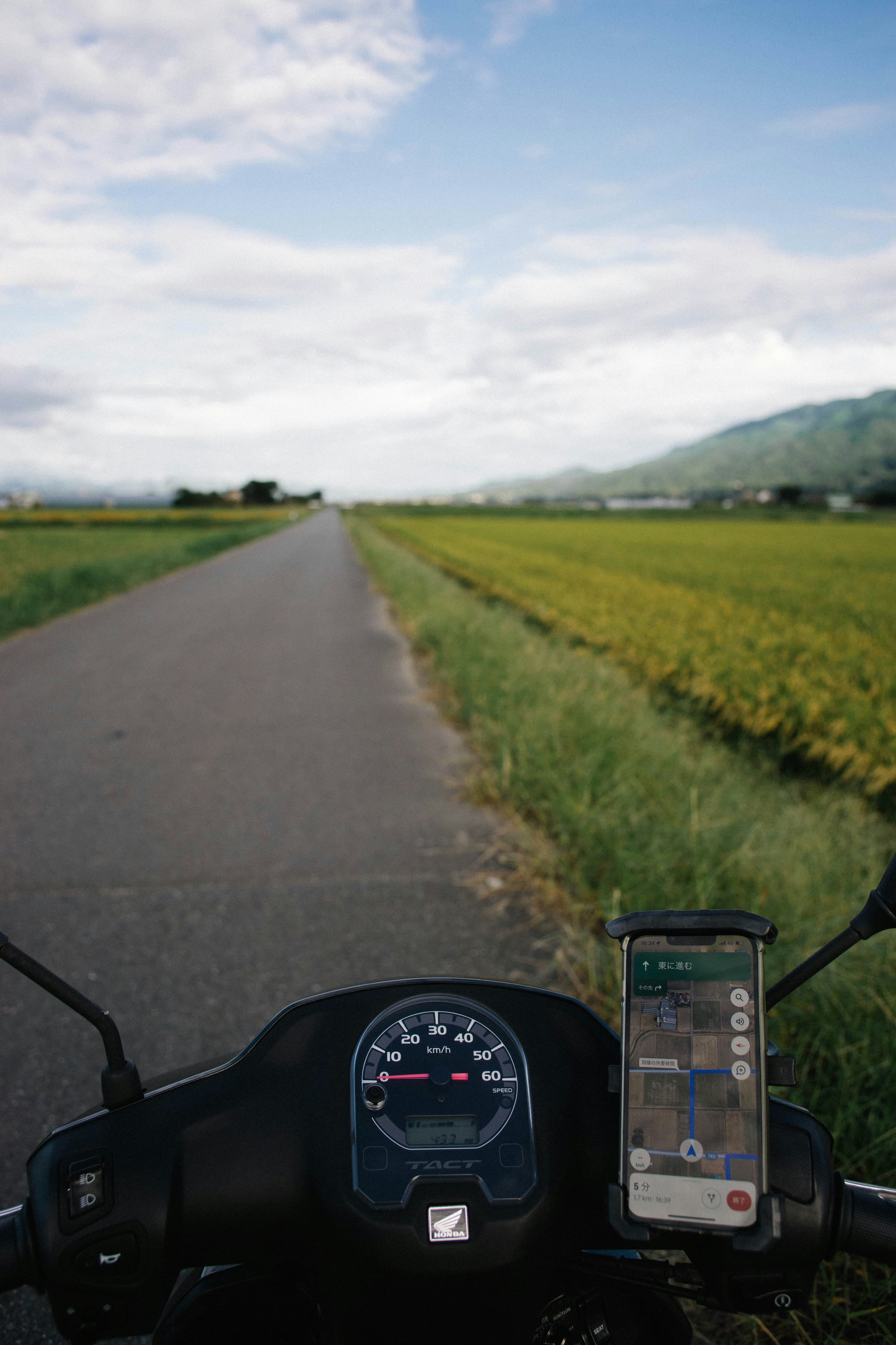 A rural Japanese country road stretching toward distant green mountains, rice fields on either side, and a motorcycle parked on the roadside under a clear blue sky (Leopold Maitre via Unsplash, free to use)