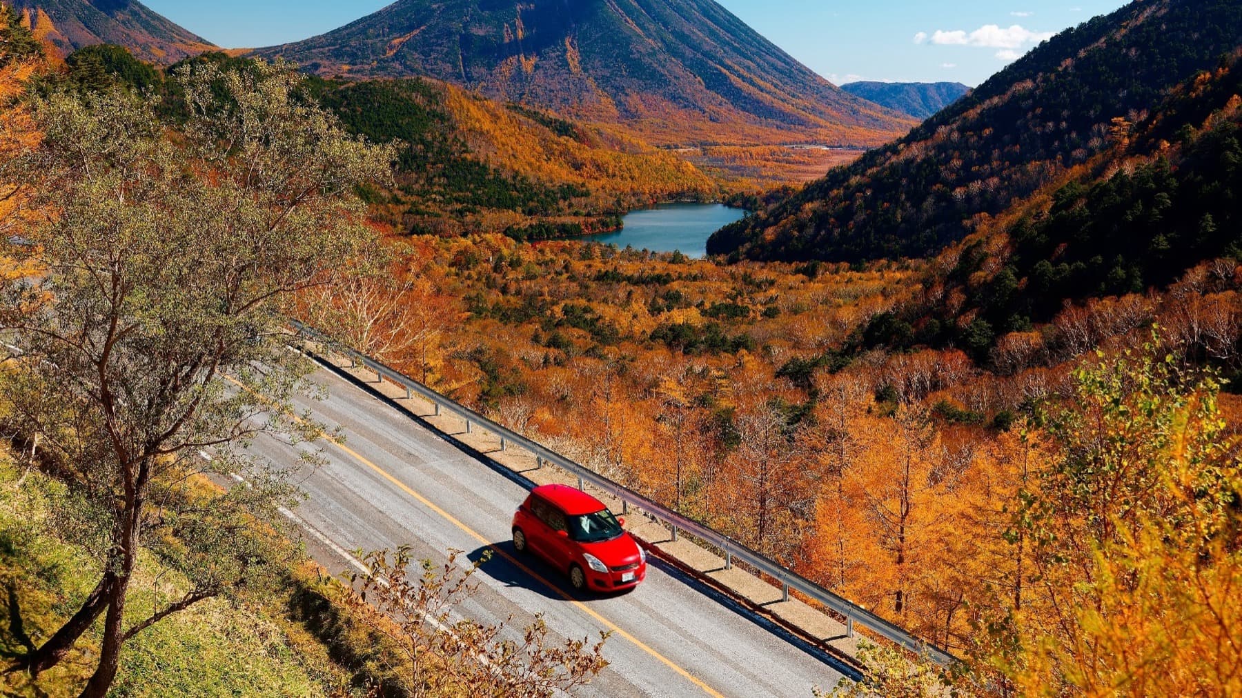 A red compact car driving on a mountain road with autumn foliage and a lake in the valley below
